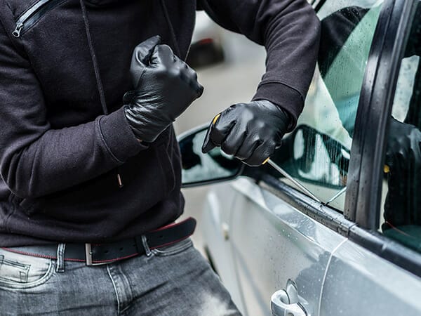 a man in a black jacket and jeans, wearing black gloves trying to break into a gray sedan with a screwdriver as part of our custom car alarm installation.